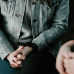 person in jean jacket with hands clasped in lap